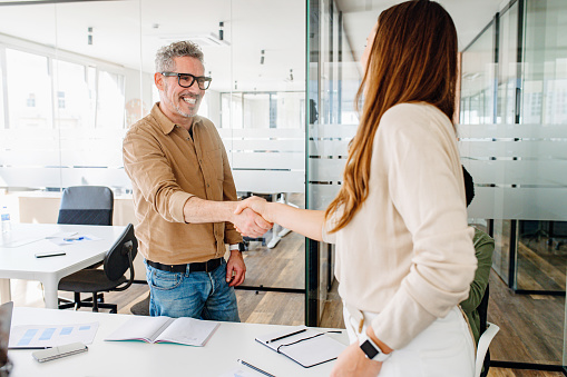 A cheerful mature man in glasses shakes hands with a female colleague in a modern office, signaling a warm welcome or successful agreement, business partners have a deal