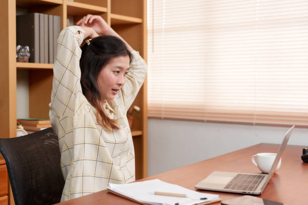 istock Asian employee stretches during a break in a home office, exhibiting a need for work-life balance. Stress relief in action, an Asian woman takes a moment to unwind at her clutter-free office desk 1987792545