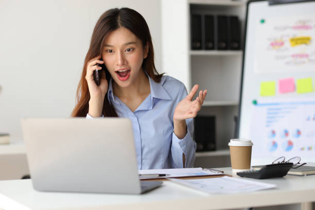 istock Asian woman in office is using mobile phone and calling someone, she is smiling with joy and surprise when receiving good news. 1987836883