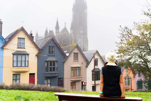 Unknown woman sitting on a bench in a park in a small town near Cork where the colorful houses and its cathedral are typical on a foggy day