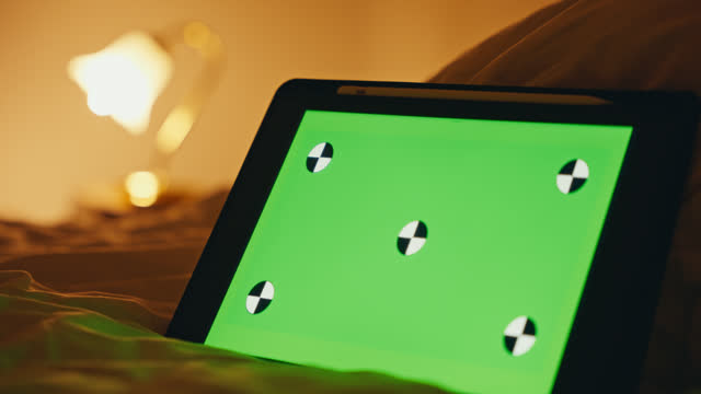 SLO MO Closeup Rack Focus of Digital Tablet with Green Empty Screen on Bed and Illuminated Lamp in Bedroom