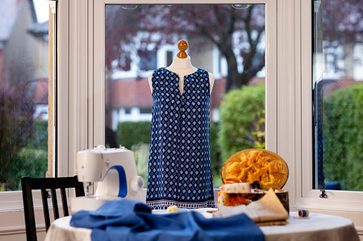 A blue patterned shift dress on a mannequin in a window in a house in Newcastle upon Tyne, England. It has been handmade by someone with great sewing skills and there is sewing equipment on the table that the mannequin is standing on.