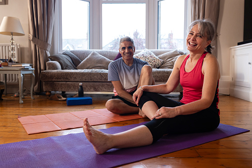 A mature couple wearing sports clothing, taking a break from meditating in their living room at home in Newcastle upon Tyne, England. They are sitting on exercise mats while looking at the camera and laughing.