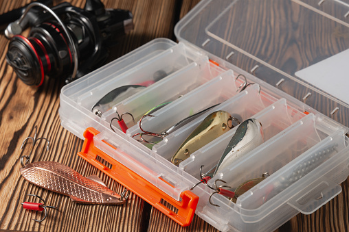 Plastic box with various metal fish baits for fishing on a wooden table.