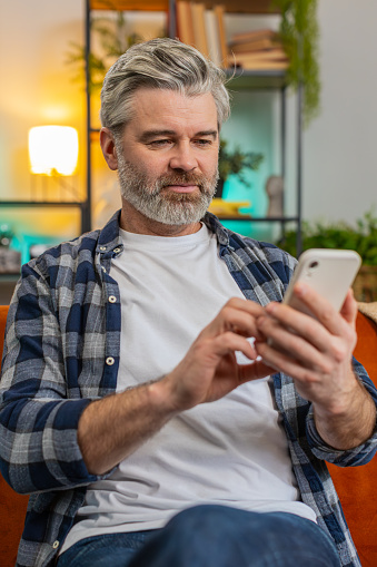 Happy mature man sitting on sofa at home getting message from old friend using smartphone look aside with smile. Grey-haired old man reading good news on phone screen in living room at home. Vertical