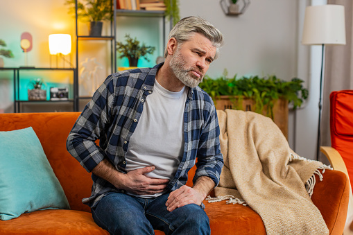 Mature ill sick man sitting on sofa feeling sudden strong abdominal stomach ache, gastritis problem. Middle-aged guy having symptom poisoning diarrhea indigestion peptic ulcer pancreatitis at home