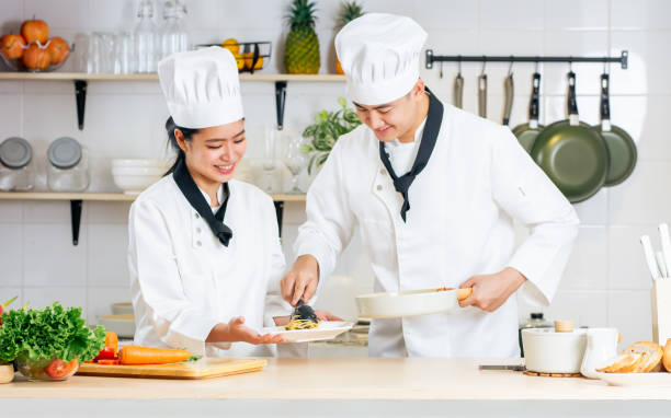istock Two Asian professional couple chef wearing white uniform and hat, helping for preparing ingredients for healthy meal, cooking in kitchen together, smiling with happiness and confidence. Food Concept. 1987913711