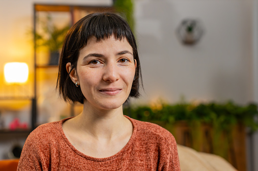 Portrait of smiling young Hispanic woman with short brown hair looking at camera. Happy content girl in casual clothes relaxing feeling satisfied concept sitting on sofa in living room at home.