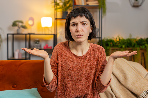 Confused woman raising hands asking why, what reason of failure, demonstrating disbelief irritation by troubles, shrugs. Caucasian girl sitting on sofa looking at camera in living room at home.