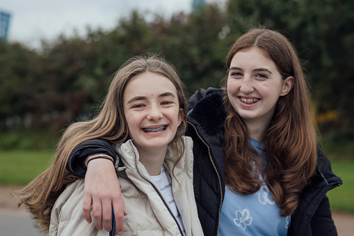A medium close up of two young teenage girls stood outside of a dental practice. They have their arm around each other and are smiling and showing off their fixed retainer braces.