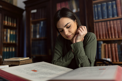 Young woman reading big old book while sitting in library in front of bookshelves. She wears green shirt and looks beautiful