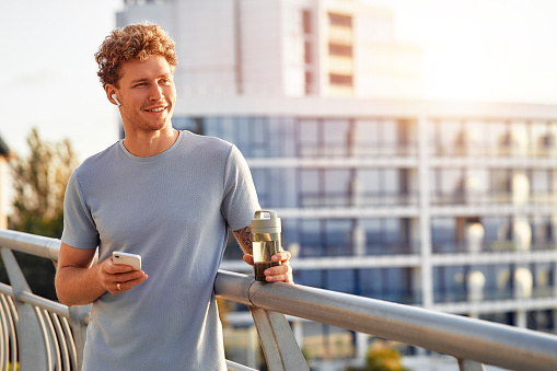 A man drinks water and uses a smartphone while resting after running outdoors on a bridge. Morning exercises and jogging. Sports and recreation, active lifestyle.