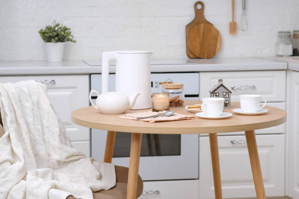istock Home spring easter interior in kitchen. Beige chair with white cover, white electric kettle, hot teapot, vase with spring branches candle, cups of tea on round wooden table in cosy white kitchen. 1987947514