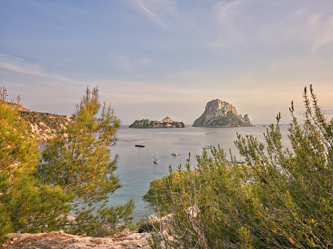 Wide-angle view of the bay of Cala d'Hort, located on the southern coast of Ibiza, renowned for its crystal-clear waters and its scenic view on the iconic rocky isles of Es Vedrà and Es Vedranell. The delicate warm light of a Mediterranean summer sunset, picturesque clouds, colourful cliffs covered with lush pine trees and Mediterranean scrub, sailing boats rocking gently on the water. High level of detail, natural rendition, realistic feel. Developed from RAW.