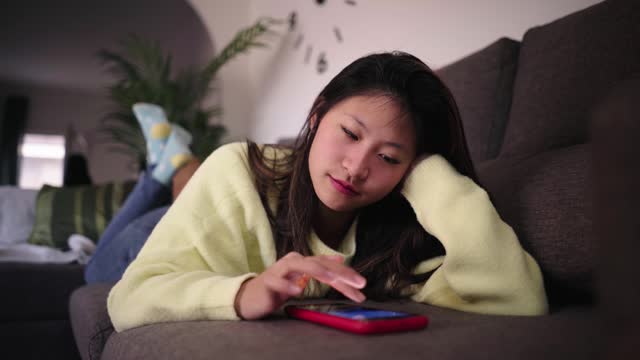 Young Asian female looking bored at cell phone lying on sofa in living room. Serious Chinese woman.