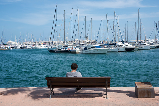 Community of Valencia, Spain - April 20, 2018: A young man sitting on a bench contemplates sailboats in the sports center of the city of Alicante