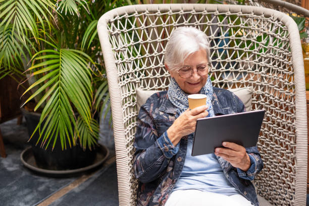 portrait of modern senior woman using digital tablet sitting on armchair with a coffee cup in hand. old generation people and new technologies - old armchair women senior adult ストックフォトと画像