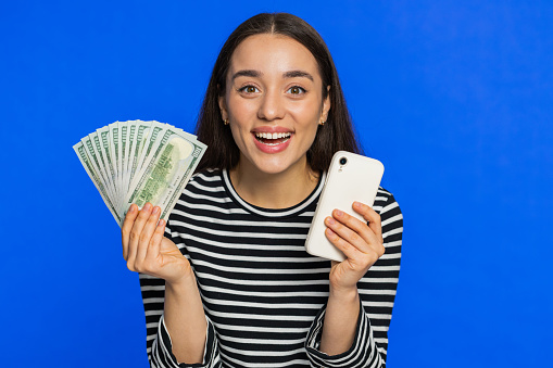Young woman looking at camera sincerely rejoicing smartphone win, receiving money dollar cash banknotes, celebrating success lottery luck. Girl isolated alone on blue studio background. Lifestyles