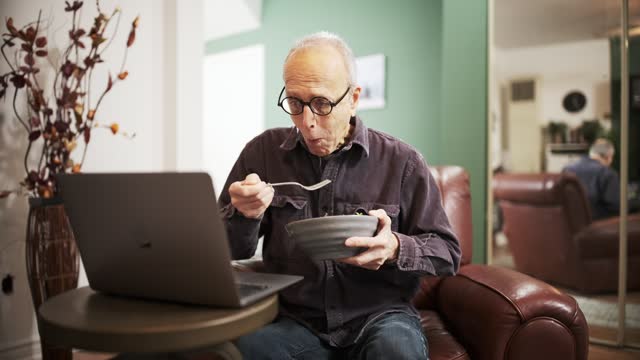 Senior man having a salad meal while using laptop in his living room