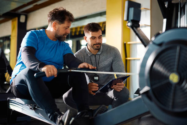 istock Young coach and mature athlete going through exercise plan during workout on rowing machine in a gym. 1988013568