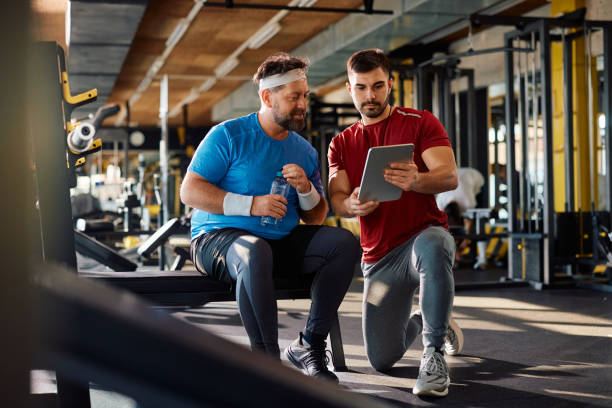 istock Fitness instructor using touchpad while showing exercise plan to mature athlete in a gym. 1988015157