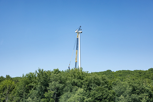 A extremely tall construction crane has lifted a large piece of the propeller hub section to the top of this extremely high, as yet incomplete, sustainable energy wind turbine electricity generator.