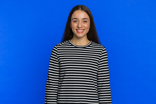 Portrait of happy Caucasian young woman in striped blouse smiling friendly, glad expression looking at camera dreaming, resting, relaxation feel satisfied good news. Girl indoors on blue background
