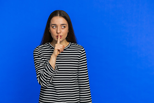 Shh be quiet please. Caucasian woman presses index finger to lips makes silence gesture sign do not tells secret stop talk gossip confidential privacy. Pretty young girl on blue background. Copy-space