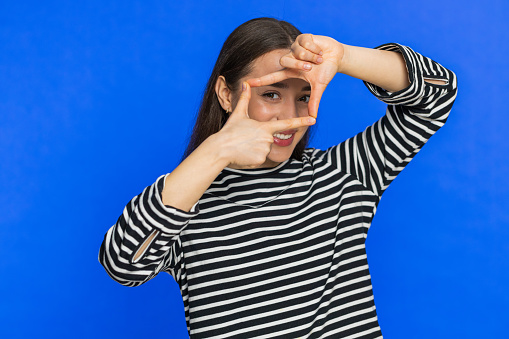 Young Caucasian woman photographer gesturing picture frame with hands looks through fingers and focusing on interesting moment imitating zoom and cropping nice image. Girl isolated on blue background