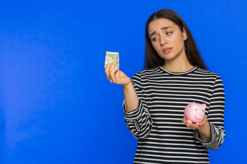 Poor Caucasian woman insufficient amount of money, holding piggybank and one dollar banknote. Financial crisis. Bankruptcy. Poverty and destitution. Girl isolated on blue background. Copy-space