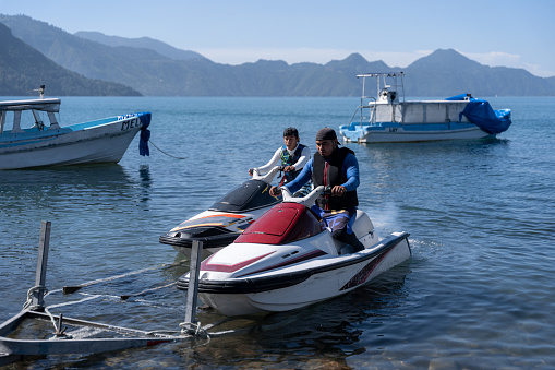 Young Hispanic men carefully lower jet skis from a trailer into the water, engaging in the process of launching the watercraft for an upcoming aquatic adventure
