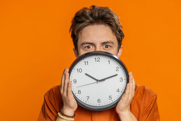 istock Serious strict young man holding office clock hiding behind checking time on watch obscuring face 1988057222