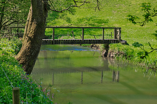 Dovedale, Peak District, Derbyshire, England, UK - Peaceful scene at a wooden bridge reflected in the River Dove in Wolfscote Dale. It is part of a beautiful walk along the footpath between Hartington and Dovedale