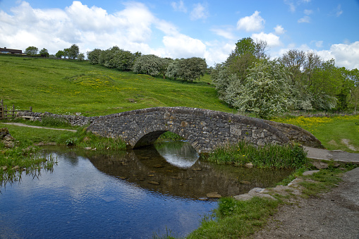 Peak District, Derbyshire, England, UK in Springtime - Old stone packhorse bridge across the River Bradford in Bradford Dale with the blue sky reflected in the water. Part of a beautiful walk, the lane crossing the bridge is part of Holywell Lane which leads to Youlgreave village