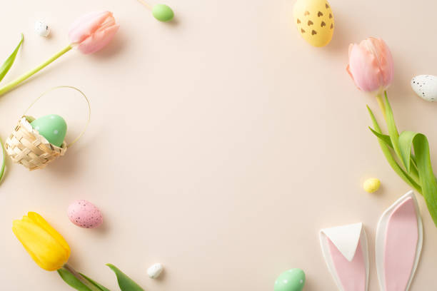 istock Springtime Joy Composition: Lively eggs, cute bunny ears, and tulips on a pastel beige background. Top view photo with an open frame ready for your text or promotional message 1988081525