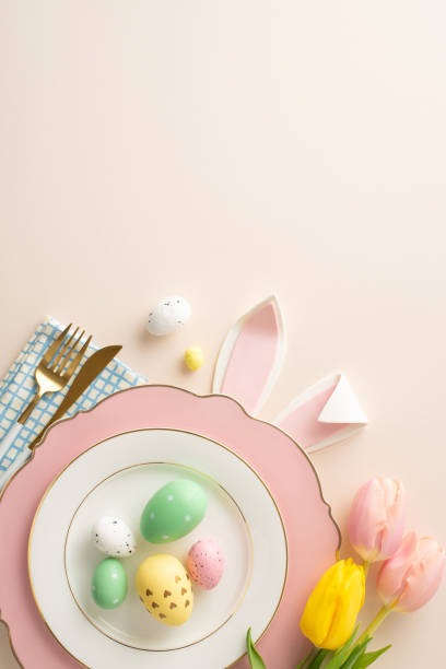 istock Easter gathering table setting vision. Top view vertical perspective of a plate filled with vivid eggs, rabbit ears, napkin, flatware, and fetching tulips on a beige background, with space for script 1988081547