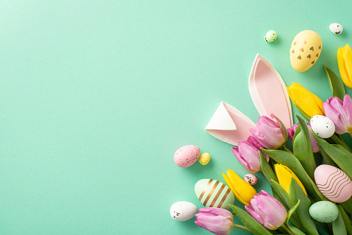 Springtime joy captured! Cheerful Easter top view scene with bouquet of vibrant eggs, playful bunny ears, and tulips. Turquoise backdrop sets the mood, leaving ample space for your festive message