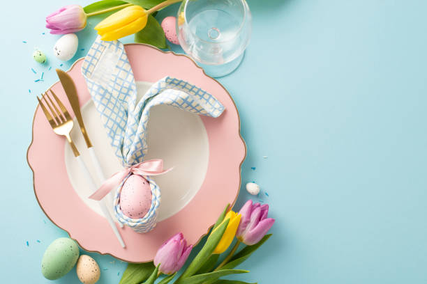 istock Easter Delight Spread: Top view photo capturing festive atmosphere with a plate, bunny ear napkin, cutlery, wine glass, tulips, sugar sprinkles, colorful eggs. Pastel blue backdrop for ad placement 1988082096