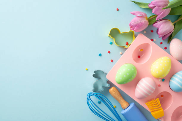 istock Easter joy in confectionery kitchen. Top view of table set for baking with whisk, roller-pin, brush. Cookie cutters, chocolate eggs in container, and more on blue backdrop. Space available for text 1988082113