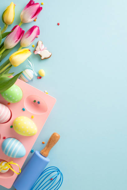 istock Easter pastry prep. A vertical top view of a baking setup with tools, adorable gingerbread, eggs in holder, sugar sprinkles, and tulips. Pastel blue backdrop, open for text 1988082564