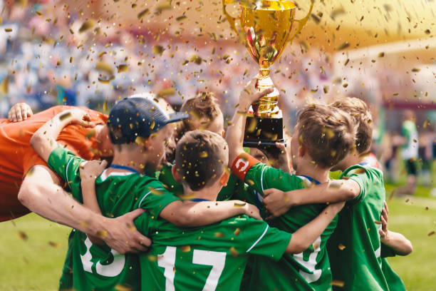 Happy children in sports team celebrating victory. Kids winning the game and rising golden cup. Kids football team huddling in circle and jumping in happiness. Youth sports team succeed in tournament niños felices en el equipo deportivo celebrando la victoria. los niños ganan el juego y levantan la copa dorada. el equipo de fútbol de los niños se acurruca en círculo y salta de felicidad. el equipo deportivo juvenil triunfa en el torneo