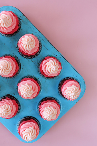 Stock photo showing close-up, elevated view of a turquoise, muffin tray of freshly baked, homemade, red velvet cupcakes in paper cake cases. The cup cakes have been decorated with swirls of ombre effect pink piped icing.  Valentine's Day and romance concept.