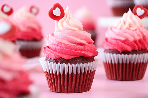 Stock photo showing close-up view of a batch of freshly baked, homemade, red velvet cupcakes in paper cake cases. The cup cakes have been decorated with swirls of ombre effect pink piped icing and sugar hearts.  Valentine's Day and romance concept.