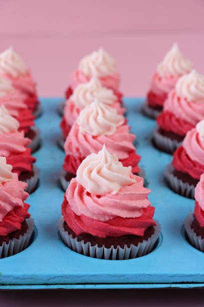 image en gros plan de turquoise, plaque de cuisson à muffins avec des rangées de cupcakes en velours rouge faits maison dans des caisses à gâteaux en papier, décorées de tourbillons pipés au beurre rose effet ombre, fond rose, mise au point sur le p - valentines day nobody plaque studio shot photos et images de collection