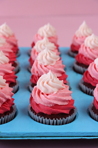 Stock photo showing close-up, elevated view of a turquoise, muffin tray of freshly baked, homemade, red velvet cupcakes in paper cake cases. The cup cakes have been decorated with swirls of ombre effect pink piped icing.  Valentine's Day and romance concept.