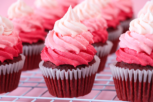 Stock photo showing close-up view of a cooling rack of freshly baked, homemade, red velvet cupcakes in paper cake cases. The cup cakes have been decorated with swirls of ombre effect pink piped icing.  Valentine's Day and romance concept.