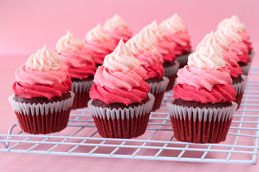 Stock photo showing close-up view of a cooling rack of freshly baked, homemade, red velvet cupcakes in paper cake cases. The cup cakes have been decorated with swirls of ombre effect pink piped icing.  Valentine's Day and romance concept.
