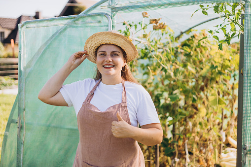 Happy 30s Woman standing in greenhouse and showing thumb up. Happy worker growing vegetables and proud of her work in hothouse. Organic farm. Agriculture concept.