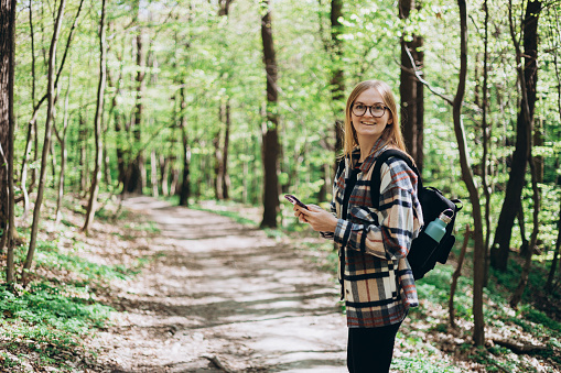 Young happy woman with backpack using smartphone and walking in the forest. Spring time. 30s Women in sport clothes hiking on woodland. Wanderlust travel concept
