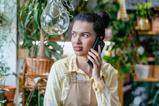 Smiling successful young female plant store owner talking with client by phone taking order standing amongst her plants in pots. Small business, entrepreneur and professions concept.